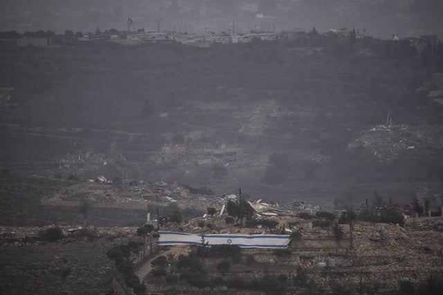 En israelisk flagga har vecklats ut vid byggnader som har rivits i en by i gränsområdena i södra Libanon. Byggnaderna på höjden i bakgrunden är en av FN-styrkans Unifils baser. Bilden är tagen i måndags.