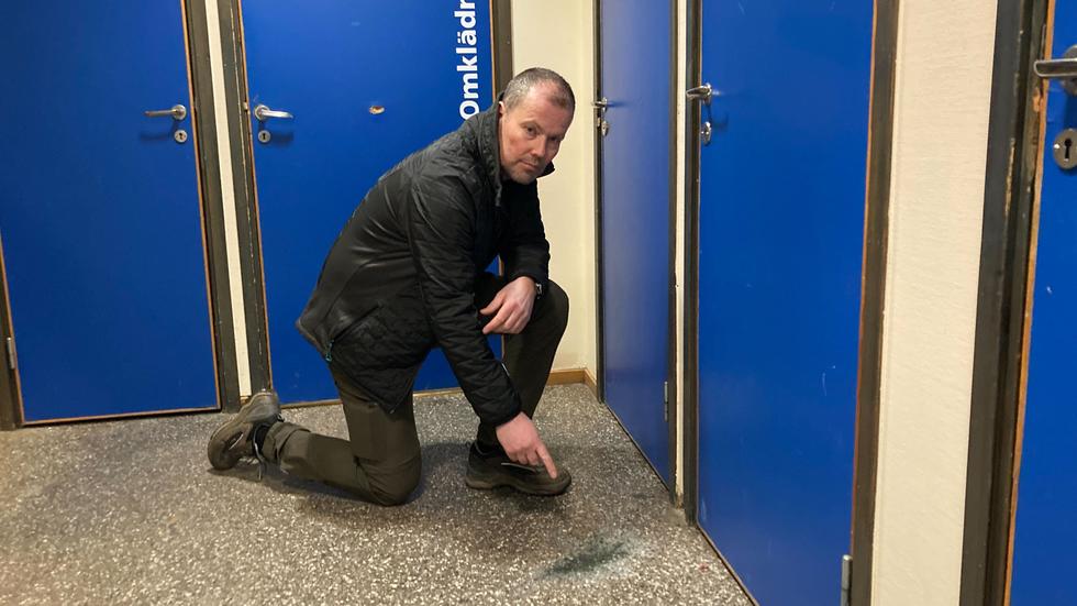 Håkan Elengård pekar på den plats där raketen lämnat ett märke i golvet. Bakom honom är dörren till omklädningsrummet där det var folk när raketen brann av. 