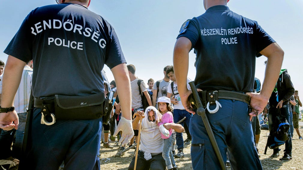Augusti 2015: Flyktingar i Ungern i väntan på vidare transport till ett läger där de ska registreras. Polis ser till att ingen lämnar uppsamlingsplatsen.